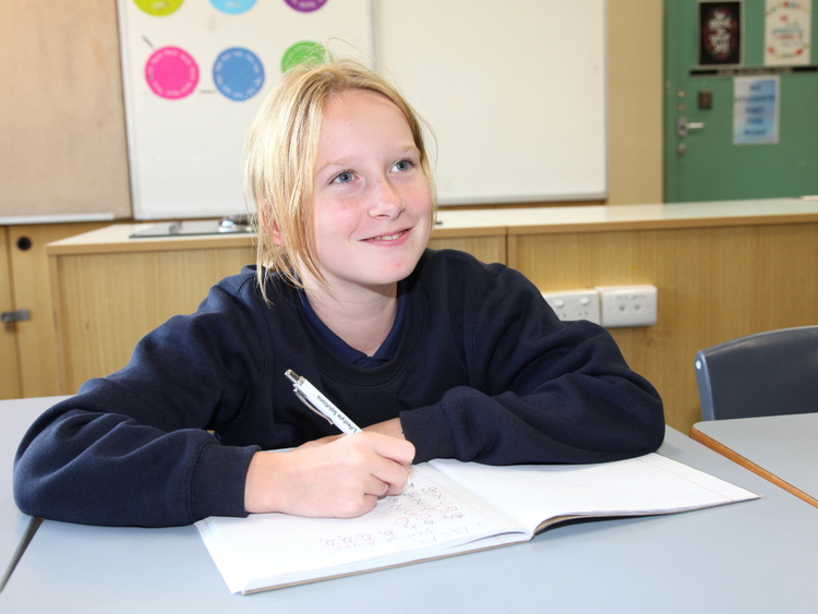 A female student in a classroom writing in a book with a pen and smiling